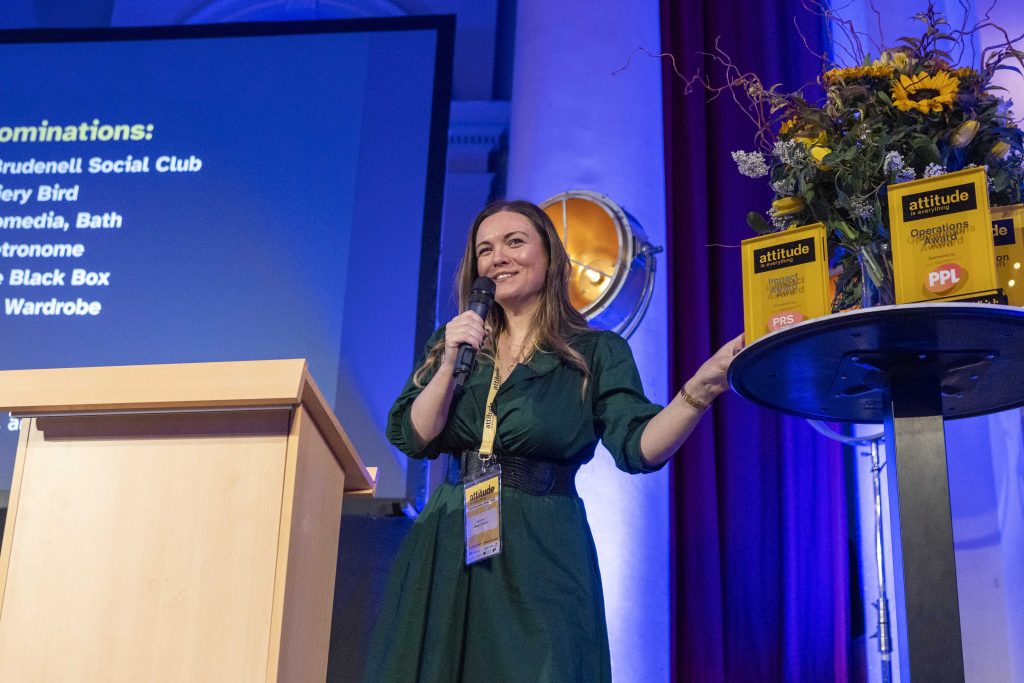 Attitude is Everything’s Interim Live Events Access Manager, Alex Covell, wears green dress, staff lanyard, and speaks into microphone. She stands by a table with awards and flowers. By her side is a podium and behind her is a screen which is projecting a slideshow.