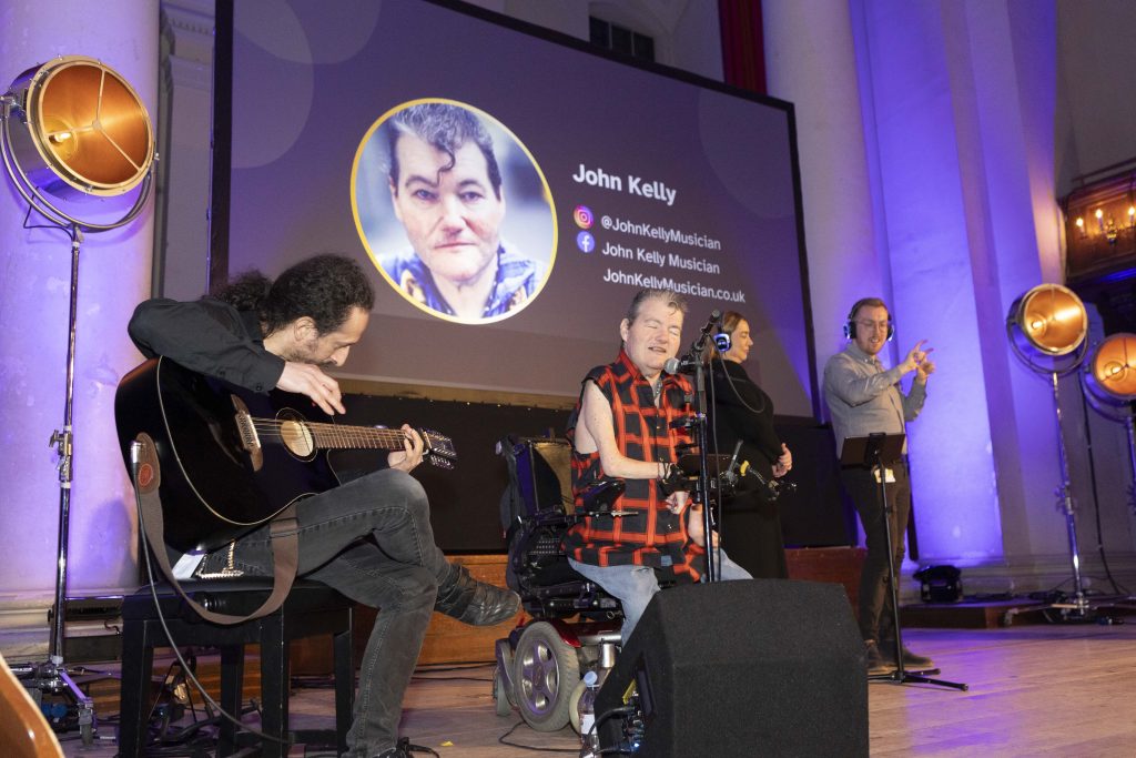 Artist John Kelly in motorised wheelchair performs on stage with his seated guitarist. To their right are 2 BSL interpreters. Behind, the screen shows John Kelly’s headshot, and social media handles: @JohnKellyMusician on Instagram, and John Kelly Musician on Facebook. His website is JohnKellyMusician.co.uk