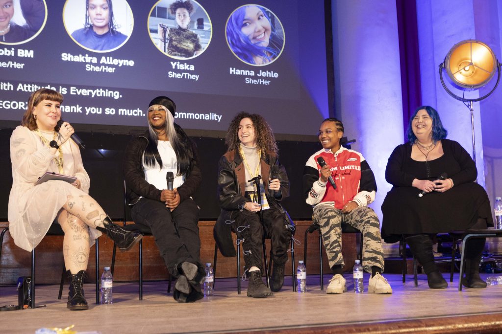 Seated panel talk on stage includes Attitude is Everything’s Inclusive Communities Manager, Bee Grzegorzek who moderates and is speaking into microphone. To their right is Nyrobi BM, Shakira Alleyne, Yiska, and Hanna Jehle. They’re all smiling at Bee!